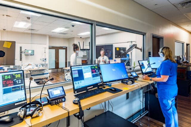 A photograph of a person working with technology that is lined up along windows which look into a nursing classroom. The classroom has two people in it, equipment, and hospital beds. One bed has a visible mannequin in it.