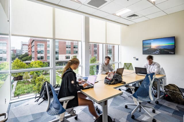A photograph of three people working on laptops at a table in a small meeting room. There are floor-to-ceiling windows letting in ample daylight. A TV is on the wall.