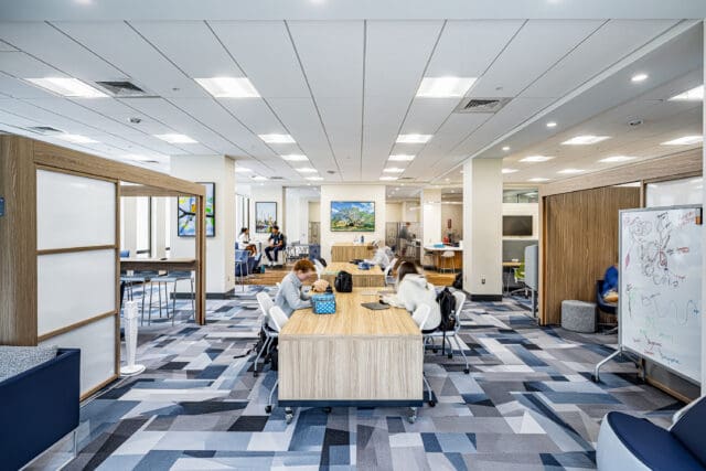 A photograph of a large common area with tables and chairs. Students are seated throughout. There are free-standing breakout spaces on each side. The carpet is a bold pattern.