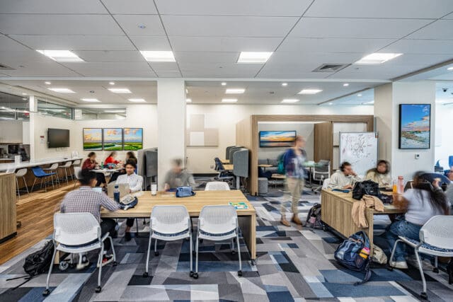 A photograph of a large common area with tables and chairs. Students are seated throughout. There are free-standing breakout spaces. The carpet is a bold pattern.