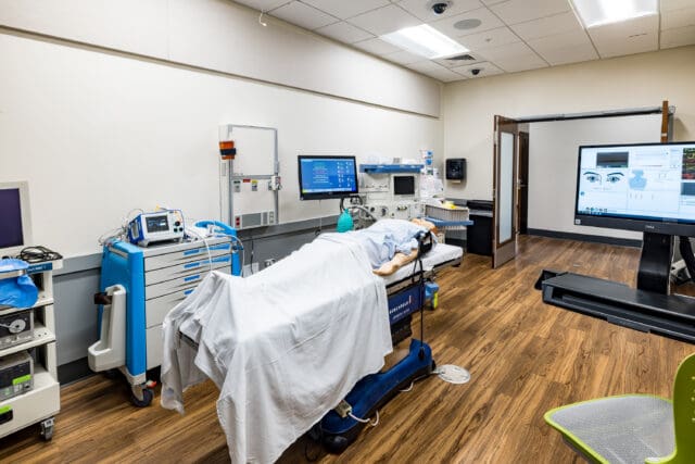 A photograph of a hospital room. A mannequin is in the hospital bed surrounded by medical equipment.
