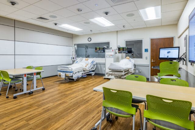 A photograph of a hospital setting with two hospital beds occupied by mannequins. A table with chairs is in the foreground.