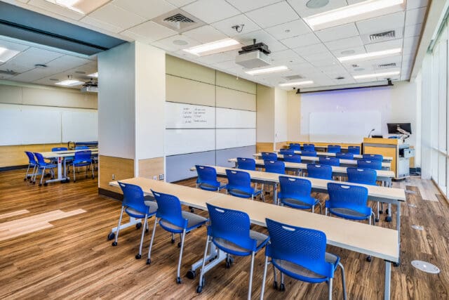 A photograph of multiple rows of desks and chairs in a classroom setting. There are multiple white boards. Additional classroom space can be seen in the background.