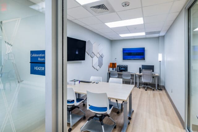 A photograph of a small office space with two tables and chairs, two televisions, and a 3D printer.
