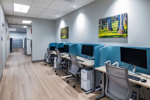A photograph of four computer workstations against a wall, each with a partition. Wall art hangs above them.