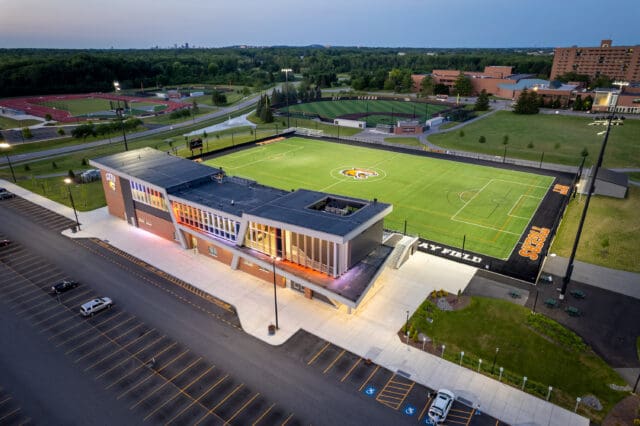 An aerial photograph of Judson Stadium and athletic field with baseball and softball fields in the distance. The stadium is lit in multiple colors. It is dusk.