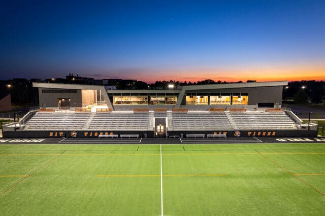 A photograph of a lacrosse field with a tiger logo at center field and a stadium in the background. It is dusk.