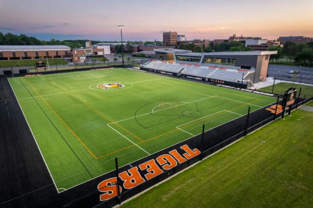 A photograph of a lacrosse field with a tiger logo at center field and a stadium and bleachers in the background. It is dusk.