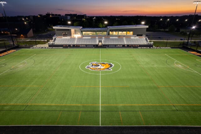 A photograph of a lacrosse field with a tiger logo at center field and a stadium in the background. It is dusk.
