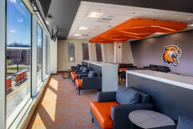 A photograph of a hospitality suite at Judson Stadium. There are lounge chairs with side tables, a unique ceiling feature, and a tiger logo on the wall. The chairs are facing a wall of windows.