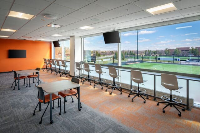 A photograph of a room with tables and chairs facing a wall of windows that overlooks a soccer/lacrosse field.