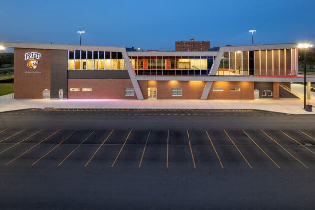 A photograph from the parking lot of a stadium with a Rochester Institute of Technology logo on it. The building is lit in many colors. it is dusk.