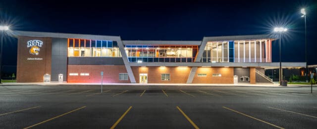 A photograph from the parking lot of a stadium with a Rochester Institute of Technology logo on it. The building is lit in many colors. it is dusk.