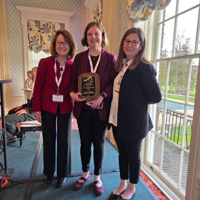 A photograph of three people standing. The person in the center is holding an award.