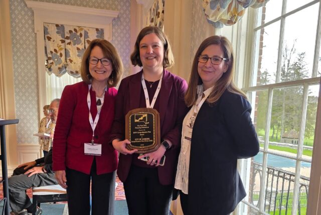 A photograph of three people standing. The person in the center is holding an award.