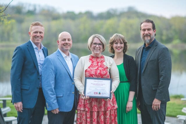 A photograph of five people standing together. The person in the middle is holding an award.