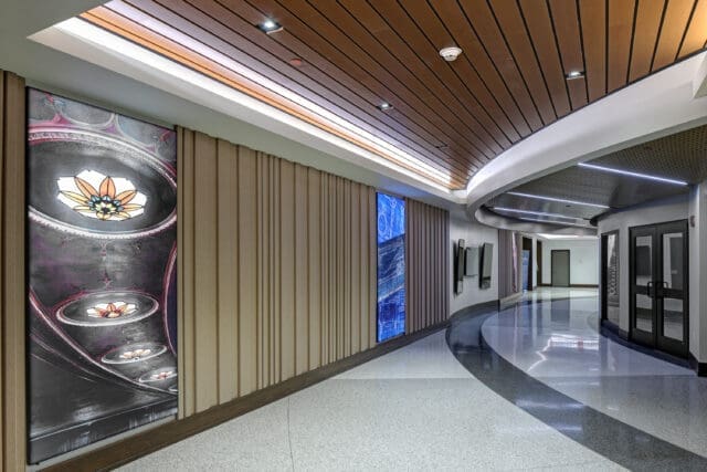 A photograph of a hallway with tall images, television screens, wood accents, and unique lighting.