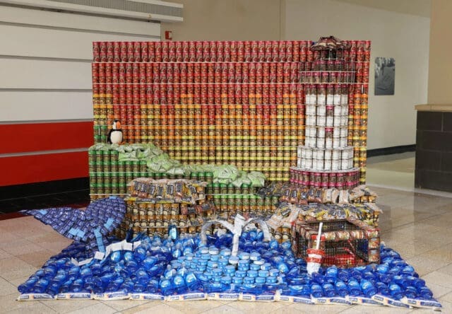 A photograph of a structure made out of canned vegetables, rice, and dish soap. It looks like a lighthouse in front of a sunset on a coast with a whale in the water and lobster trap above the water.