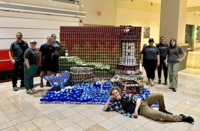 A photograph of five people around a structure made out of canned vegetables, rice, and dish soap. It looks like a lighthouse in front of a sunset on a coast with a whale in the water and lobster trap above the water.