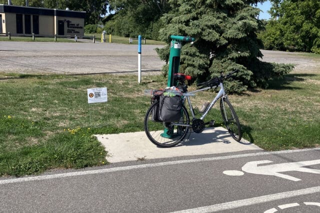 A photograph of a bicycle sitting up against a pole along a walk/bike path. A small yard sign is visible.