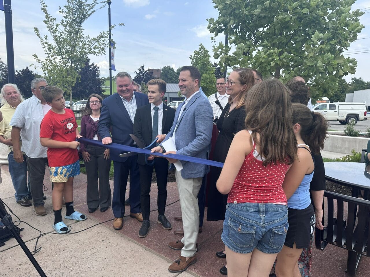 A photograph of a group of people gathered in an outdoor park area for a ribbon-cutting ceremony. People are holding a ribbon and the person in the center has a large pair of scissors.