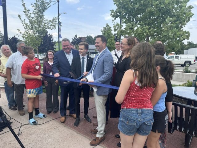 A photograph of a group of people gathered in an outdoor park area for a ribbon-cutting ceremony. People are holding a ribbon and the person in the center has a large pair of scissors.