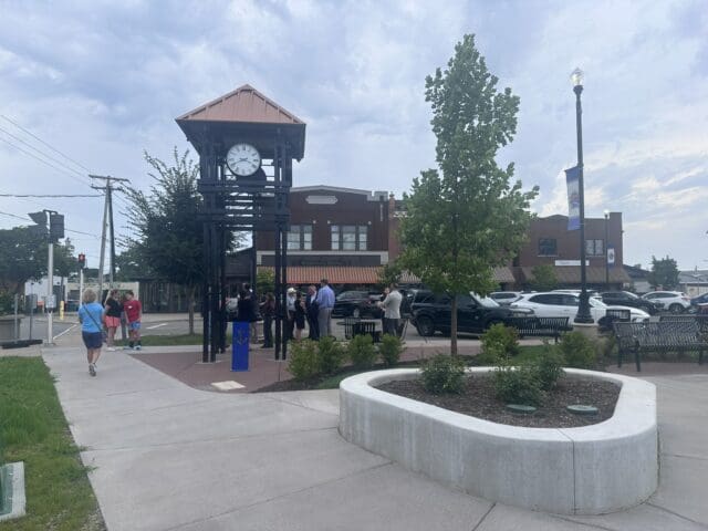 A photograph of a small park with a clock tower feature, shrubs, trees, and a walking path. People are gathered in the background.