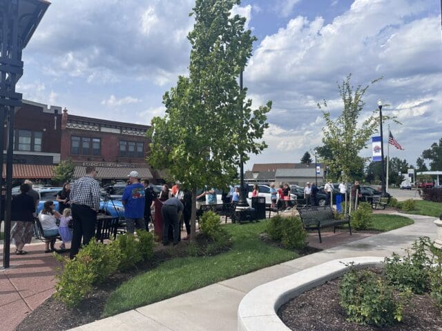 A photograph of a small park with shrubs, trees, and walking paths. People are gathered in the background.