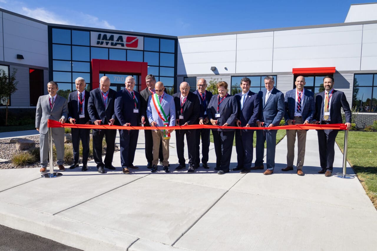 A photograph of a ribbon-cutting ceremony. A group of people are standing in a line holding a red ribbon, each with a pair of scissors. They are in front of a building with a sign that says IMA.