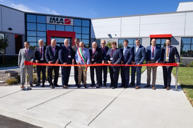 A photograph of a ribbon-cutting ceremony. A group of people are standing in a line holding a red ribbon, each with a pair of scissors. They are in front of a building with a sign that says IMA.
