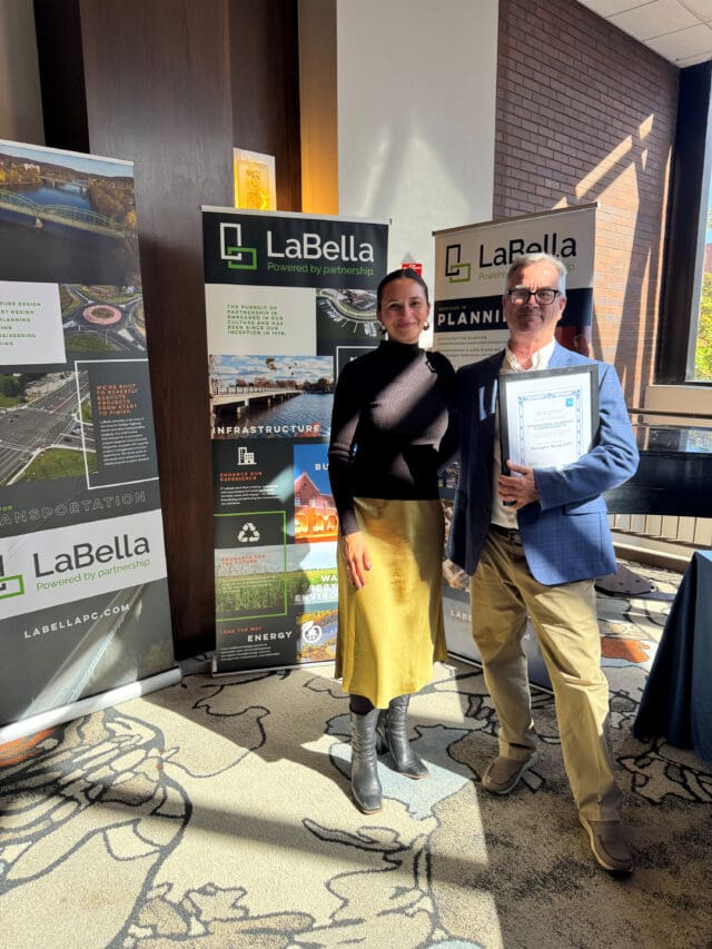 A photograph of two people standing in front of three banners for LaBella Associates. One person is holding an award.